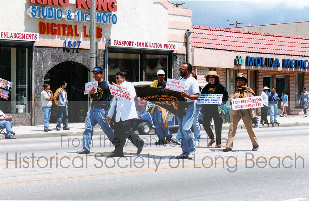 Viradet David Kreng (center in white jacket) walks the parade route alongside a representative from California State University, Long Beach. They hold signs in Khmer and English with messages like "Knowledge comes from education - Rewards come from discovery."