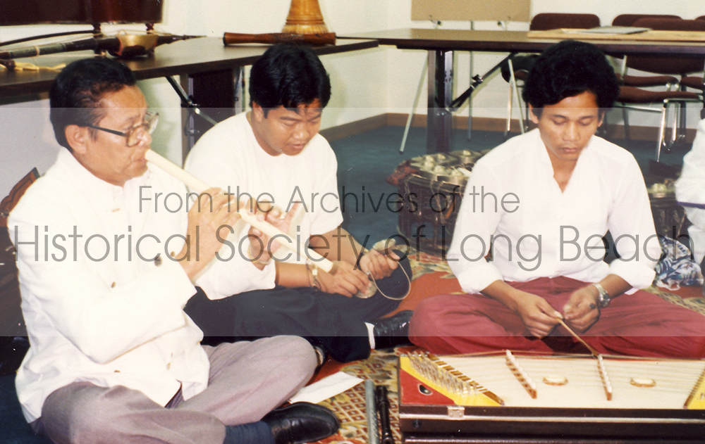 Master musician Yinn Pon plays the flute (left) while musician strikes small hand cymbals (center) and the other instrument prepares to pluck the Khim, a flat, trapezoidal-shaped string instrument encased in a wooden body with metal strings stretched across its surface.