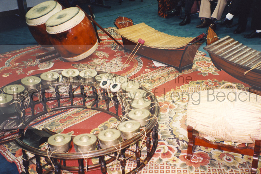 Photo of the following Cambodian traditional instruments from right to left: Cambodian drum, Skor (front right); Cambodian xylophone-like instrument, Kong Vong Thom (left front); Pair of Cambodian large drums, Skor Thom (left back); Pair of Cambodian gong percussion, Roneat Ek (left right).