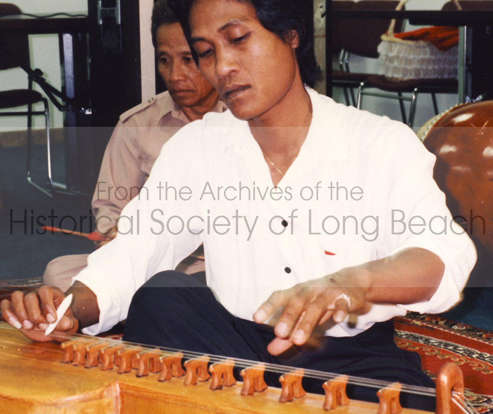 Close-up image of a musician playing the Cambodian string instrument called Takei. The Takei is a long wooden instrument with 4 strings that the musician plucks.