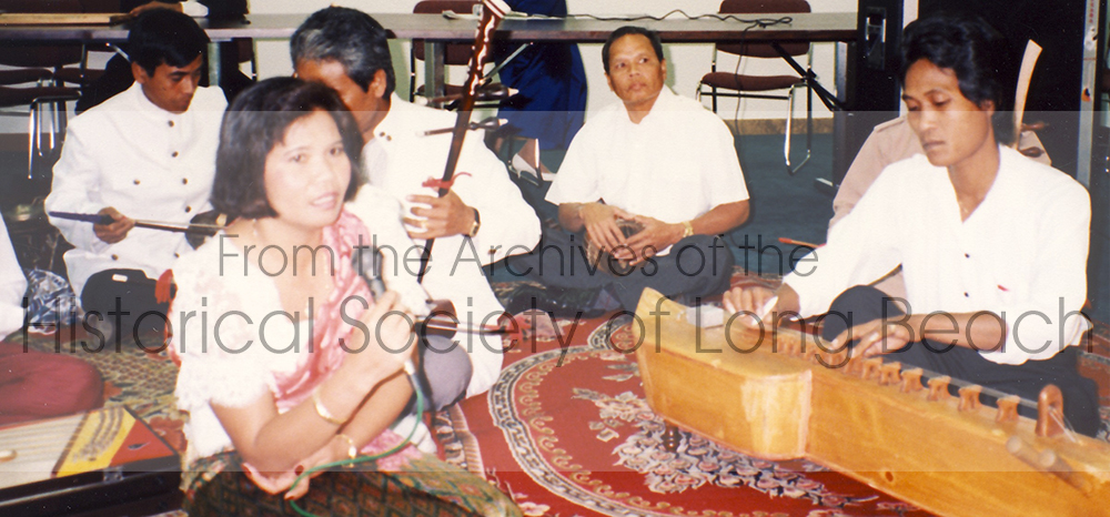 Image of musicians playing (from left to right): Cambodian woman sings into microphone while musicians playing the Tro Ou, a 2-3 string instrument played with a bow, and another musician plucks the Takei (front right).