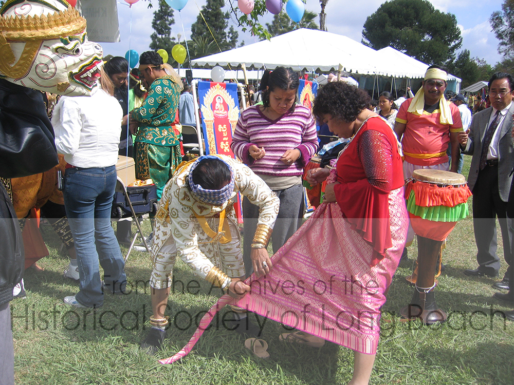 Performers help each other get dressed for their performances at Cambodian New Year in Long Beach. The woman is being helped with her kbun. An 8’ length of silk is wrapped about her waist toward the front. The length of cloth is being twisted and will be pulled to her back between her legs. It will be held in place with a belt.