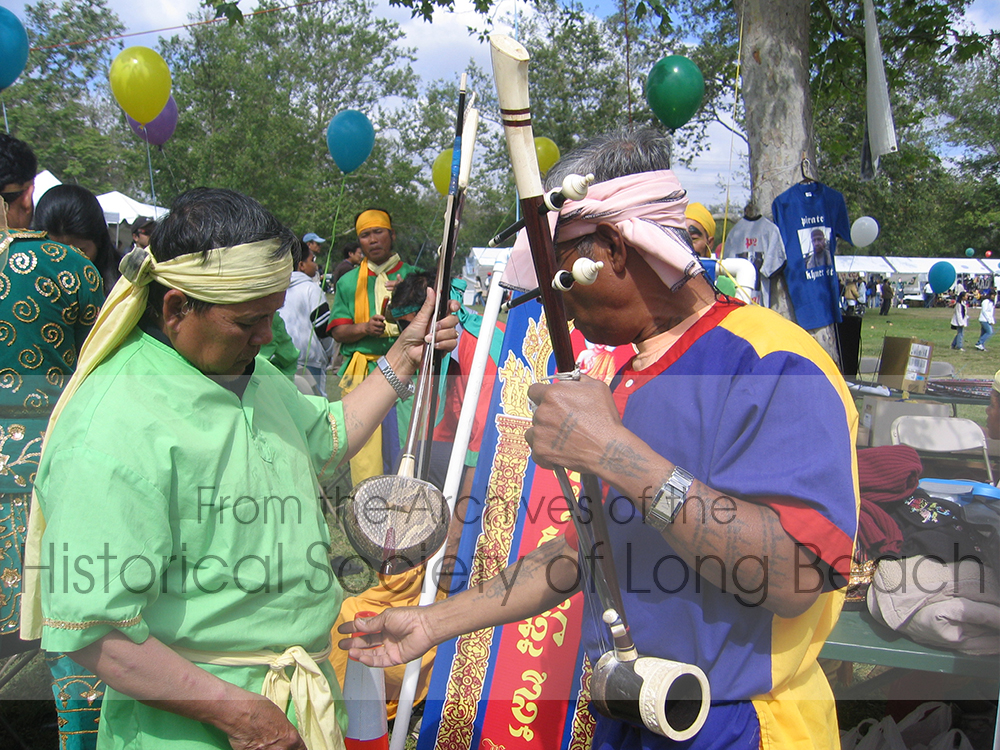 Two Chhayam performers with their bowed, string instruments, called Tro. The sound box of the Tro can be made from a variety of materials. The man in green is holding a Tro made of coconut shell with a snake skin head. The man in blue and yellow is holding a Tro made of blackwood and ivory with metal trim.