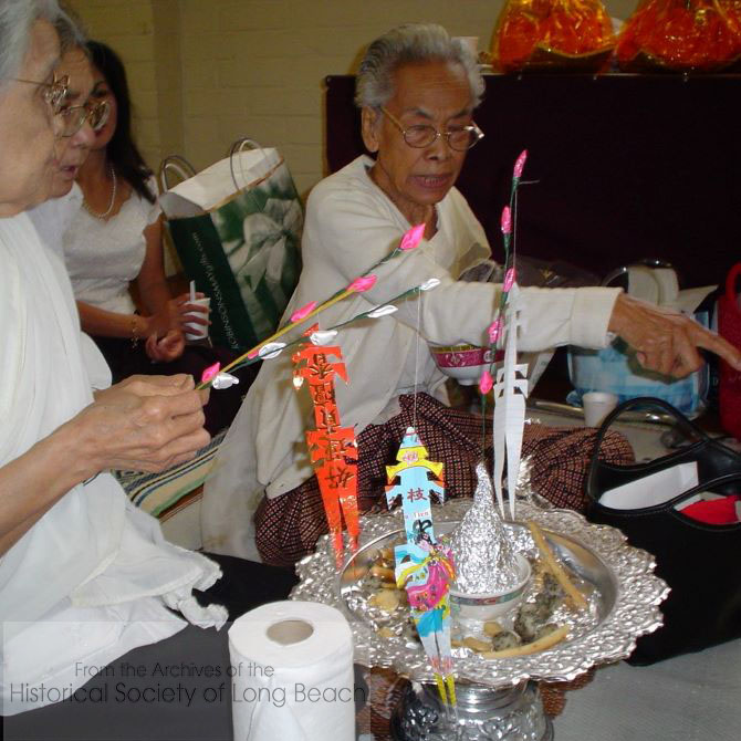 2003 Pchum Ben ceremony at Wat Willow (Khemara Buddhikaram). A group of women have gathered to prepare special offerings. They are wearing white to indicate they have taken vows to follow the five Buddhist precepts for a period of time. The five precepts are: do not kill, do not steal, do not engage in sexual misconduct, do not lie, and do not drink intoxicants. The orange, multicolored, and white "flags" on the silver platter are small versions of the much larger flags hung outside a house during a funeral.