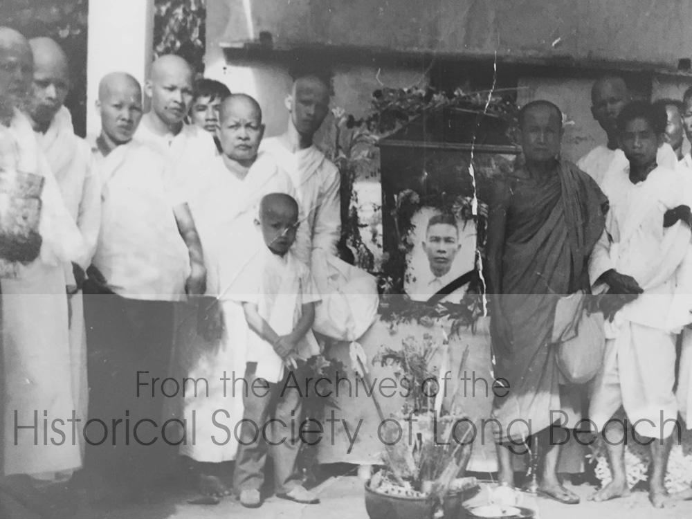 Funeral of Clark Tang’s grandfather, Cheark Ung, at a temple in Phnom Penh, Cambodia 1974. Cheark Ung’s photo and coffin are at center. Back row (left to right): Kim Ung, uncle; Pang Ung, uncle; Kim Heang Ung, uncle. Middle row (left to right): You Long, grandmother. Clark is the child in front. People to the right are unidentified. The grandfather died of a stroke or heart attack before the war. Family members shaved their heads and wore white out of respect for the deceased. Kim Ung had been a military policeman and his entire family was killed during the Khmer Rouge period. Kim Heang Ung was a doctor and was also killed during the Khmer Rouge.