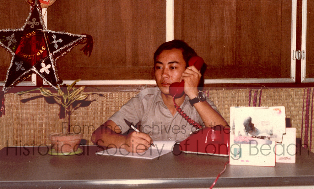 Dan Durke studied hard to become proficient in English. He was hired to work for the Catholic Relief Services office in their nutrition department. He is pictured here talking on the phone with the UNHCR (United Nations High Commissioner for Refugees) office at the transit center in the Khao-I-Dang refugee camp in Thailand, c. 1982. The star behind him is a common Christmas decoration.