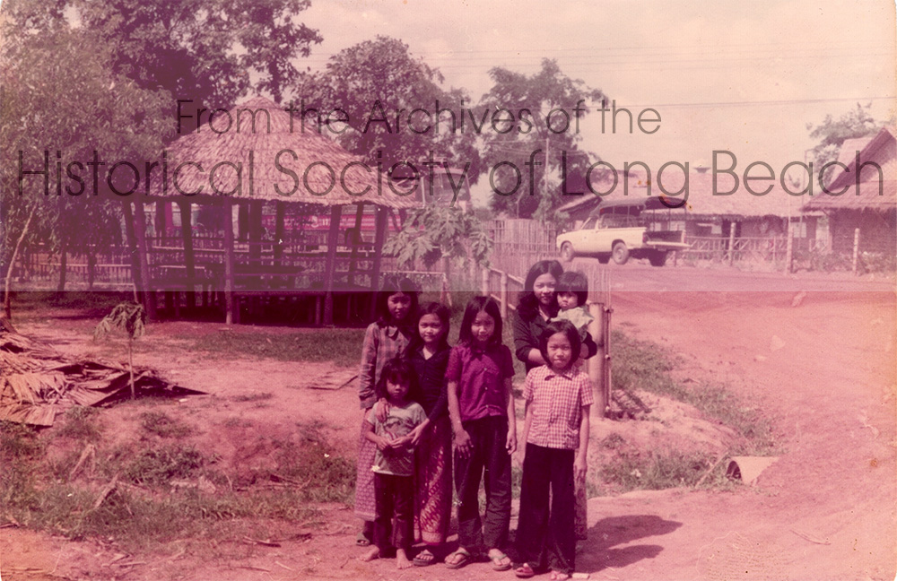 Kam Lou Lopez’s family at a holding facility (“prison”) before being accepted into the Khao-I-Dang refugee camp, 1981. Kam is in the back on the right holding a baby. Her sister, Meicy, is on the left in the black shirt. The other children are unidentified.