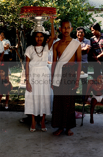 Monorom Neth’s ordination ceremony to become a novice monk, at the first Cambodian Buddhist temple, located in Lakewood, CA, in 1985. The ordination ceremony occurs in stages. At this stage, Monorom’s hair and eyebrows have been shaved to help eliminate vanity. His sister, Somealea, is walking with him to the main sanctuary where he will be ordained. She carries his robes on a silver tray. The robes are protected by an embroidered and beaded covering called “trai.” His other sister, Somealy, will also take a turn carrying the robes. The next stage will be to appear before the elder monks who will lead Monorom through the ceremony and from whom he will receive blessings and his robes. Following this, he will discard his old clothes, change into his robes, and return to the sanctuary as a novice monk. He will receive final blessings and acceptance from the monks, as well as well wishes from those in attendance.