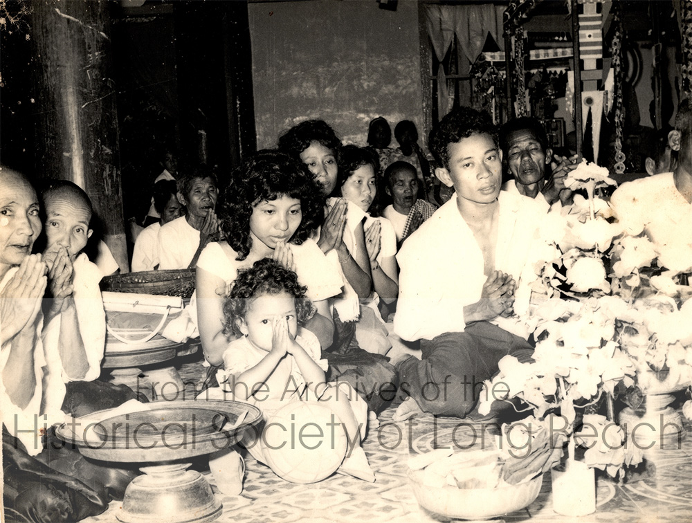 Namoch Sokhom’s family before his birth at a fundraising event at one of the Buddhist temples his father, Luy Sokhom (in foreground, looking at the camera), helped to build in Kampong Cham Hanchey, Cambodia, 1963. At far left are Namoch’s two great aunts. Next to Luy is Namoch’s mother, Thach In with Namoch’s older sister, Namom, sitting in front of her. Luy’s cousin, Su An, is behind them. Behind Luy to his right is Namoch’s maternal grandfather, Pho. Luy Sokhom was an accomplished architect and artist, who built many temples.