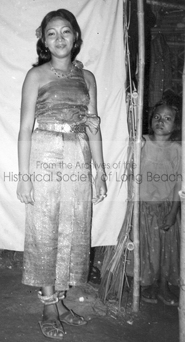 Black and white photograph of a young woman dressed in Cambodian dance attire standing in front of a screen. A small child on the right peeks from behind the screen.