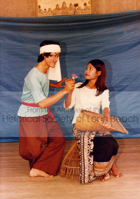 Two people on a stage in front of a blue screen. They are dressed in Cambodian folk attire. The male (left) is handing a flower to the young woman (right) who is seated on a fishing trap.