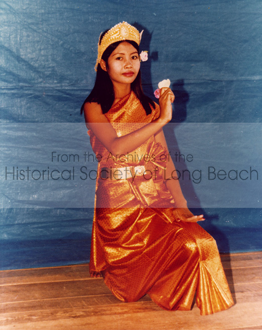 Young woman is dressed in Cambodian dance attire in front of a blue screen. She is holding a flower.