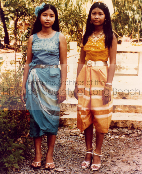 Two young women dressed in Cambodian dance attire. They are outside. The woman on the left is dressed in blue. The other is dressed in gold.